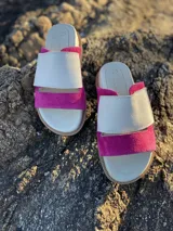 Sandalias bajas de cuero blanco con dos tiras: una tira ancha de gamuza verde en una sandalia y rosa en la otra, y una tira fina de cuero blanco.