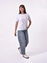 Remera de manga corta color beige claro con cuello redondo. Presenta un estampado gráfico central con un plato de pasta rodeado por un borde azul y blanco, sobre un fondo de franjas verticales rojas y blancas. El estampado incluye texto en cursiva en tonos turquesa y rojo.