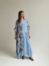 Vestido largo celeste con gran estampado floral marrón en el lateral derecho, de corte holgado, mangas 3/4 con trabilla y dobladillo irregular con flecos desflecados. Incluye un bolsillo aplicado en el frente.