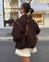 Chaqueta de jean color marrón oscuro, con diseño de hombros caídos y detalle de tachas metálicas en el cuello y la parte superior de las mangas. Presenta cierre frontal con botones metálicos.
