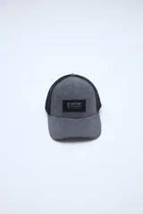 Gorra tipo trucker gris con visera curvada, paneles frontales de tela y paneles traseros de red negra. Cuenta con un parche rectangular negro con inscripción en el frente.