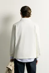 Remera polo de manga larga de corte regular fit, confeccionada en algodón color blanco crudo. Presenta cuello solapa blanco, cierre frontal con botonadura oculta y una franja horizontal ancha en el pecho en color azul y verde.