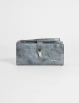 Billetera plegable de cuero vegano con acabado metalizado en tono azul grisáceo. Cuenta con una correa de mano (strap) integrada, cierre principal y un broche metálico decorativo en el frente. Incluye logo de la marca en la parte inferior.