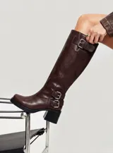 Bota de caña alta estilo biker confeccionada en cuero color marrón oscuro. Presenta tres hebillas metálicas decorativas, cierre lateral y taco cuadrado de madera.