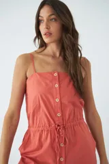 Monoprenda corta de color verde oliva, con tirantes finos ajustables, cierre frontal con botones y cintura elástica con cordón ajustable. Cuenta con bolsillos laterales.