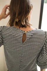 Blusa de tela con estampado vichy en blanco y negro, con cuello redondo y mangas con tres volados escalonados que llegan hasta el codo. Presenta puños elásticos y cierre con botón en la parte posterior.