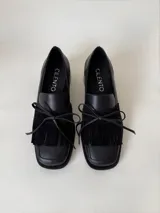 Mocasín de cuero marrón con textura granulada, diseño de horma cuadrada, detalle de flecos y lazo decorativo en el empeine, montado sobre una suela y taco bajo de color negro.