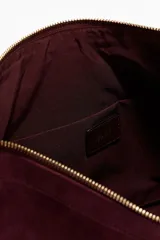 Bolso de hombro color borgoña, con textura que simula gamuza o ante, de forma curva y cierre superior con cremallera. Posee una correa de hombro delgada de cuero marrón oscuro y un tirador de cremallera con borla a juego.