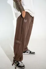 Pantalón de estilo urbano color marrón, con cintura elástica ajustable mediante cordón, bolsillos laterales y bajos ajustables con tiras. Presenta un pequeño estampado en la pierna.
