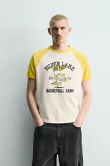 Camiseta de corte relajado con mangas raglán cortas en color amarillo y cuerpo en color crema. Presenta un estampado gráfico frontal con temática de campamento de básquetbol y un cocodrilo.