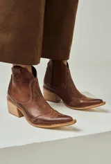 Texana de caña baja color chocolate, confeccionada en cuero con acabado envejecido. Cuenta con puntera perfilada, tacón ancho de madera y sutiles costuras decorativas en la parte superior.