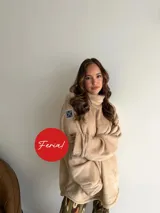 Buzo de peluche color beige, con cuello alto, mangas largas y diseño oversize. Presenta un parche decorativo en la parte superior de la manga derecha.