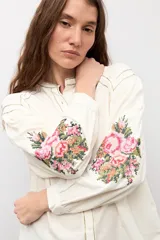 Camisa de color natural con mangas largas abullonadas y bordado floral multicolor en los puños. Presenta cuello mao y cierre frontal con botones.
