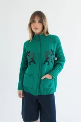 Cardigan de punto color marrón con cierre frontal, cuello alto y bolsillos aplicados. Presenta diseño tejido localizado con detalles en violeta y negro en el pecho.