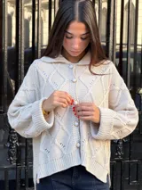 Saco tejido de punto con diseño de ochos y trenzas, color beige. Presenta una silueta oversize, cuello estilo camisero y cierre frontal con botones tipo joya. Posee mangas holgadas y un diseño de largo asimétrico.
