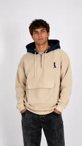 Hoodie beige con capucha azul marino, cierre frontal parcial con cremallera y bolsillo tipo canguro delantero. Presenta un pequeño bordado en el pecho.