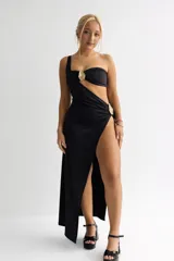 Vestido largo negro de diseño asimétrico con un solo hombro. Presenta detalles de herrajes dorados en el escote y en la cintura, con una abertura lateral profunda en la falda que aporta movimiento y estiliza la silueta.