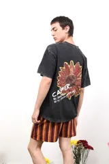 Remera de algodón con efecto lavado ácido en color negro. Presenta un diseño estampado en la espalda que incluye una ilustración de una flor en tonos rojizos y amarillos, acompañada por el texto 'CAPER' y frases motivacionales.