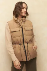 Chaleco puffer color crema, con cuello alto, cierre frontal y cuatro bolsillos con solapa. Cuenta con cintura ajustable con cordón.