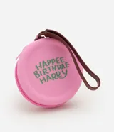 Monedero redondo de silicona color rosado con cierre de cremallera y correa de mano marrón. Presenta un estampado temático en color verde con la frase 'Happee Birthdae Harry'.