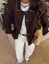 Chaqueta de jean color marrón oscuro, con diseño de hombros caídos y detalle de tachas metálicas en el cuello y la parte superior de las mangas. Presenta cierre frontal con botones metálicos.