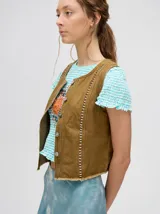 Chaleco de denim liviano color marrón, con cierre frontal de botones metálicos y detalle de tachas plateadas verticales. Presenta terminación desflecada en el ruedo.