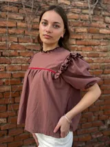 Blusa color chocolate con mangas cortas abullonadas, cuello redondo con volados y detalles de costura en zigzag de color rojo en el pecho y volados oscuros en los hombros.