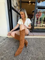 Botas texanas de caña alta en eco gamuza color marrón, con punta afilada y taco bajo. Presentan bordados decorativos en la caña.