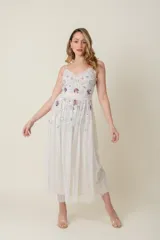 Vestido midi de tul color marfil con escote en V y tirantes finos. El corpiño y la falda están adornados con lentejuelas y apliques bordados en tonos pastel como lila, rosa y azul, simulando flores. La falda es voluminosa y con capas de tul.
