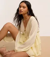 Blusa femenina de chiffon color amarillo pálido, con escote en V profundo, lazada ajustable en el centro del busto y mangas largas ajustadas. Presenta un diseño con volados en el ruedo.