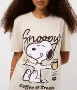 Remera de algodón color beige con cuello redondo y mangas cortas. Presenta una estampa frontal centrada del personaje Snoopy sosteniendo una taza y una dona, acompañada por el texto 'Snoopy's Coffee & Treats'.
