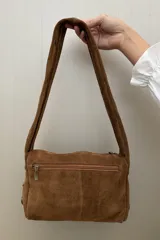 Cartera de hombro confeccionada en gamuza color habano, con diseño de correa ancha decorada con tachas metálicas circulares. Presenta cierre superior y placa metálica con logo en el frente.