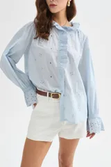 Camisa de algodón con detalles de broderie, volados y cuello a la base. Presenta diseño holgado, cierre frontal con botones y mangas largas con puños fruncidos.