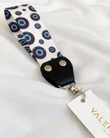 Llavero de tela blanca con estampado de ojos turcos azules y negros, conector de cuero sintético negro y gancho metálico plateado.