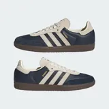 Zapatillas Adidas Samba OG con exterior de cuero azul marino y detalles en gamuza beige. Presentan las icónicas tres tiras laterales, suela de goma color marrón y el nombre del modelo en letras doradas.