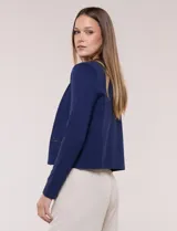 Cardigan corto azul marino de punto, con silueta entallada y largo a la cintura. Cuenta con un ribete decorativo que recorre el cuello redondo y la abertura frontal, integrando una cadena dorada en contraste. Posee bolsillos delanteros simulados y mangas largas.
