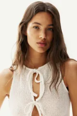 Top corto sin mangas de punto color blanco roto, con escote redondo y abertura frontal que se cierra mediante tres lazos anudados. Presenta un detalle de peplum con ligero vuelo en el bajo.