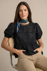 Blusa negra de algodón con corte oversize, mangas cortas abullonadas y detalles de encaje y bordados en el frente y hombros. Presenta un diseño con pliegues verticales y terminaciones con puntilla.