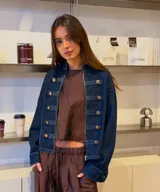 Chaqueta de denim azul oscuro con estilo militar, cuello alto, cierre frontal con cremallera y detalles de trabas decorativas con botones dorados.