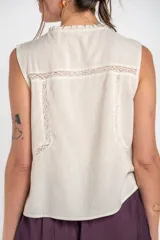 Blusa sin mangas de color beige, confeccionada en tela ligera con detalles de bordados florales frontales, encajes y cierre delantero con tiras para anudar.