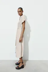 Vestido midi camisero de color beige, con cuello solapa, manga corta y cierre frontal con botones. Incluye un cinturón del mismo tejido con hebilla metálica.