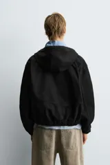 Cazadora negra de algodón con efecto lavado, corte relajado y capucha ajustable. Presenta cierre frontal de cremallera, bolsillos frontales tipo canguro y elásticos en puños y bajo.