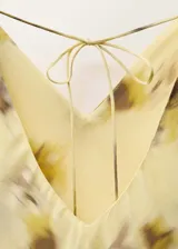 Vestido largo de tirantes finos con cuello de pico, confeccionado en viscosa con estampado floral difuminado en tonos amarillos y marrones. Presenta un diseño recto con detalle de cadena metálica a la altura de la cadera y cierre de lazo en la espalda.