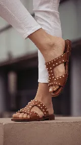 Sandalia plana de cuero color tostado con diseño de tiras anchas y tachas doradas distribuidas por toda la capellada.