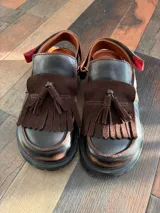 Mocasines de cuero color marrón oscuro, con diseño de flecos y tira con hebilla en el empeine, y suela plana con plataforma baja.