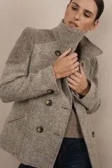 Chaqueta corta de lana color beige con diseño de espiga, doble abotonadura, cuello solapa y bolsillos ojal. Presenta apliques de tela en los hombros.