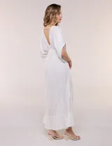 Vestido largo blanco confeccionado en viscosa con hilos de lurex, escote en V profundo con lazo y borlas, mangas kimono y falda fluida con volados en el ruedo.