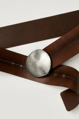 Cinturón ancho de cuero marrón oscuro con efecto desgastado y pespunteado sutil. Cuenta con una hebilla redonda grande de metal plateado envejecido, sin orificios visibles, que funciona como un nudo o broche cruzado.
