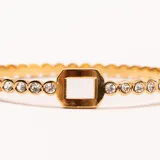 Brazalete rígido dorado con incrustaciones de cristales transparentes y un detalle central rectangular calado.