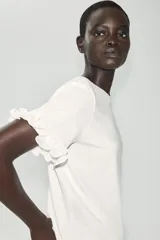 Camiseta blanca de algodón con cuello redondo, manga corta y detalle de aplicación de volantes en las mangas.